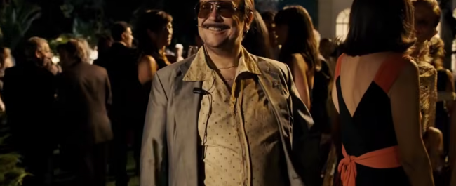 Santiago Segura caracterizado como José Luis Torrente sonríe con gafas de sol y traje desaliñado en una fiesta elegante nocturna, en un fotograma de 'Torrente, presidente'.